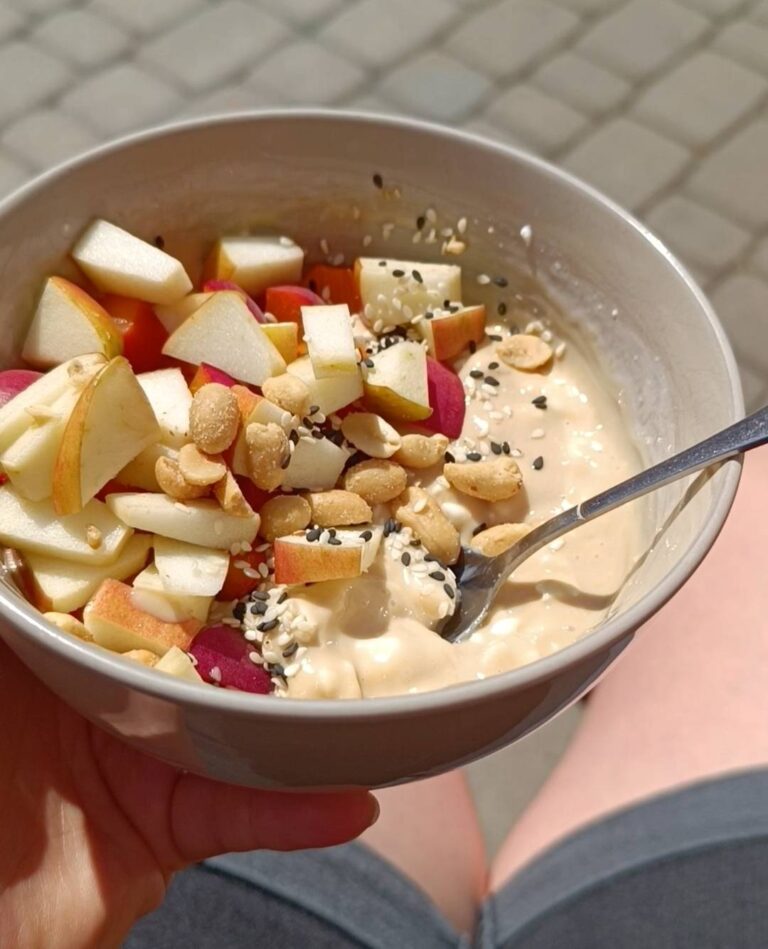 Szara miska, a w niej puddingt proteinowy wymieszany z serkiem wiejskim. Na wierzchu ułożone pokrojone jabłko i brzoskwinia, posypane sezamem i orzeszkami ziemnymi.